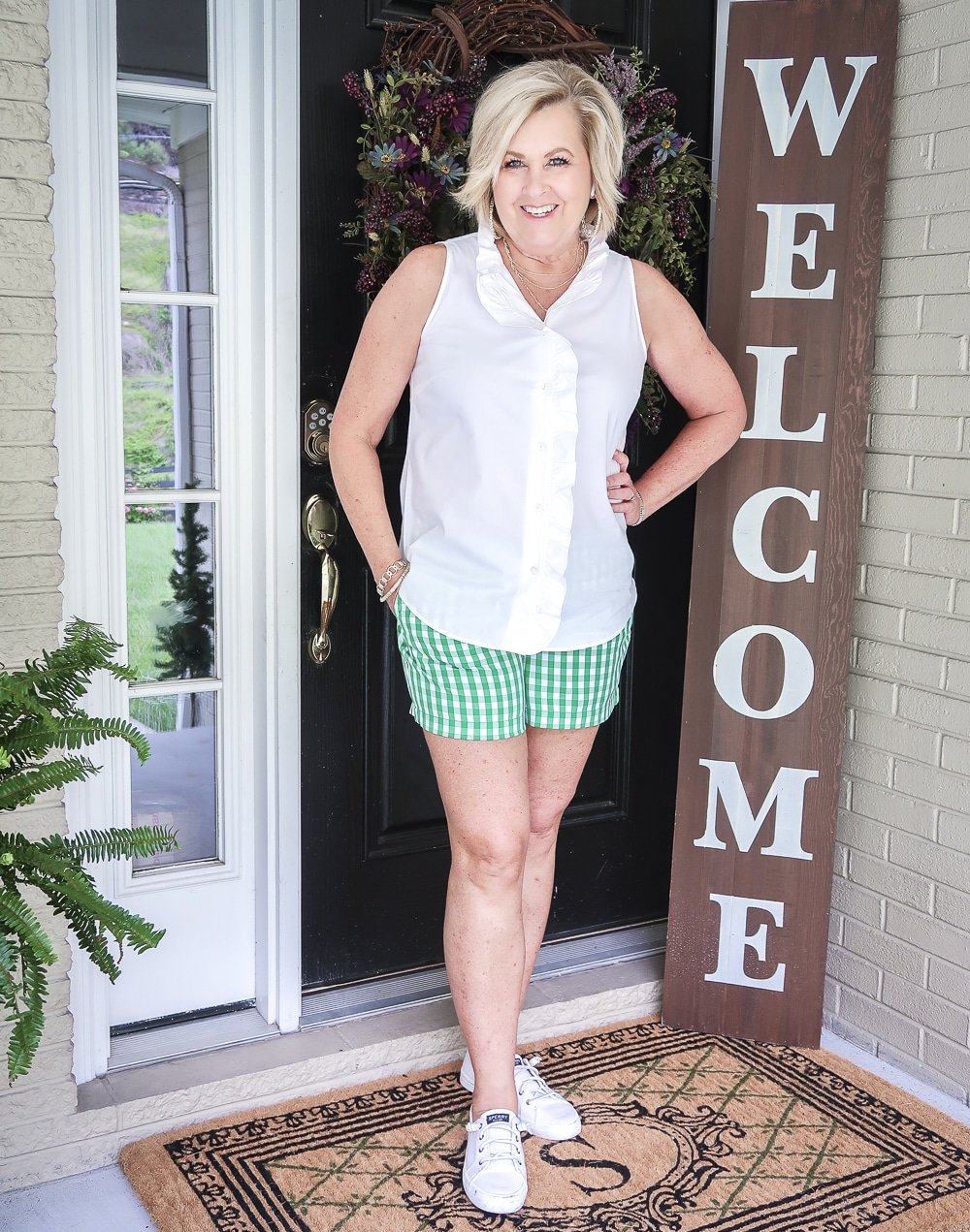 FASHION BLOGGER 50 Is Not Old wearing a sleeveless top with a ruffled placket and green gingham shorts with a pair of white slip on sneakers. 