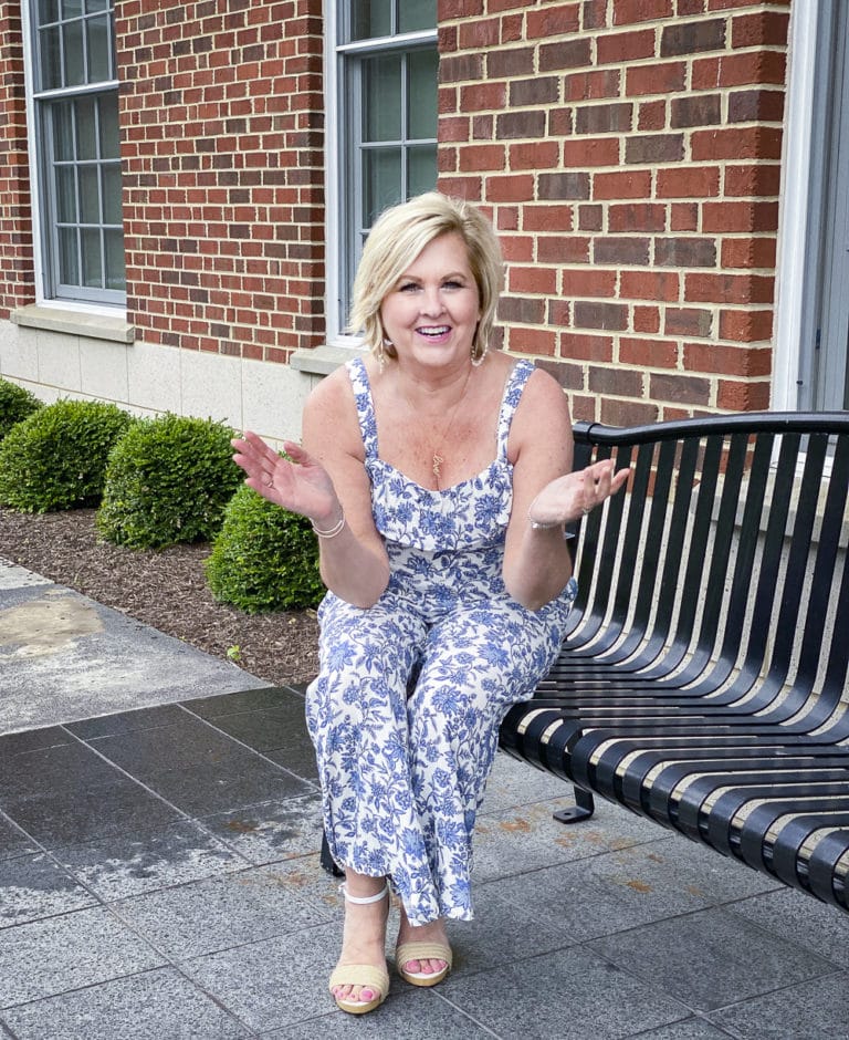 Fashion Blogger 50 Is Not Old is sitting on a bench laughing while wearing a blue and white jumpsuit from Loft with white espadrille wedge sandals