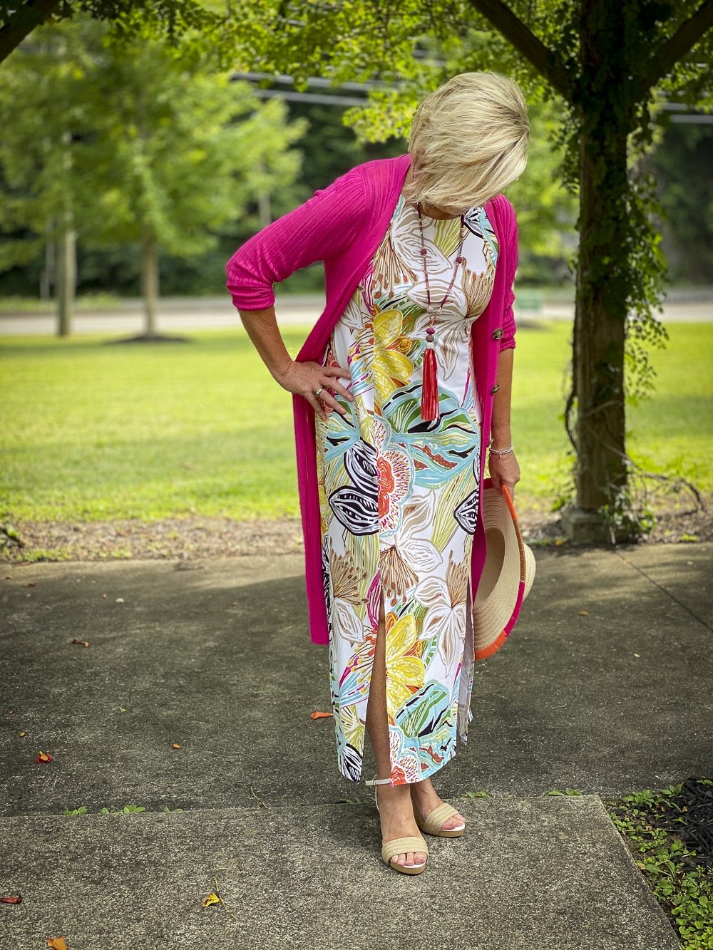 Fashion Blogger 50 Is Not Old is looking down while wearing a tropical print sleeveless dress with a pink cardigan, colorful accessories and white espadrilles