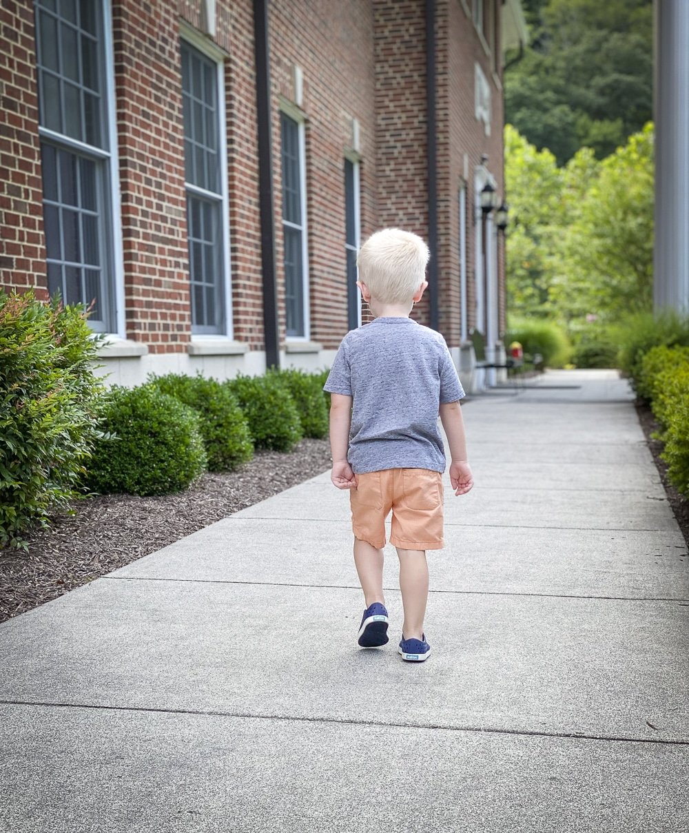 Fashion Blogger 50 Is Not Old's grandchildren in their back to school outfits. He is wearing a gray henley tee shirt, orange shorts from Carters, and navy blue Native sneakers while walking away.