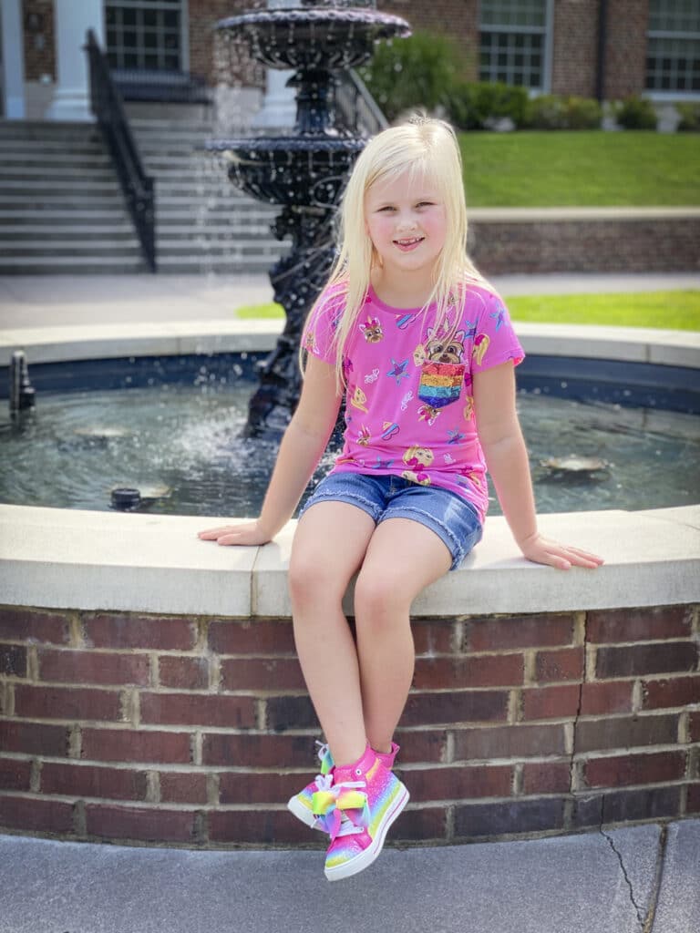 Fashion Blogger 50 Is Not Old's grandchildren in their back to school outfits. She is sitting by the water fountain in her pink JoJo Siwa top and denim shorts with rainbow colored sneakers
