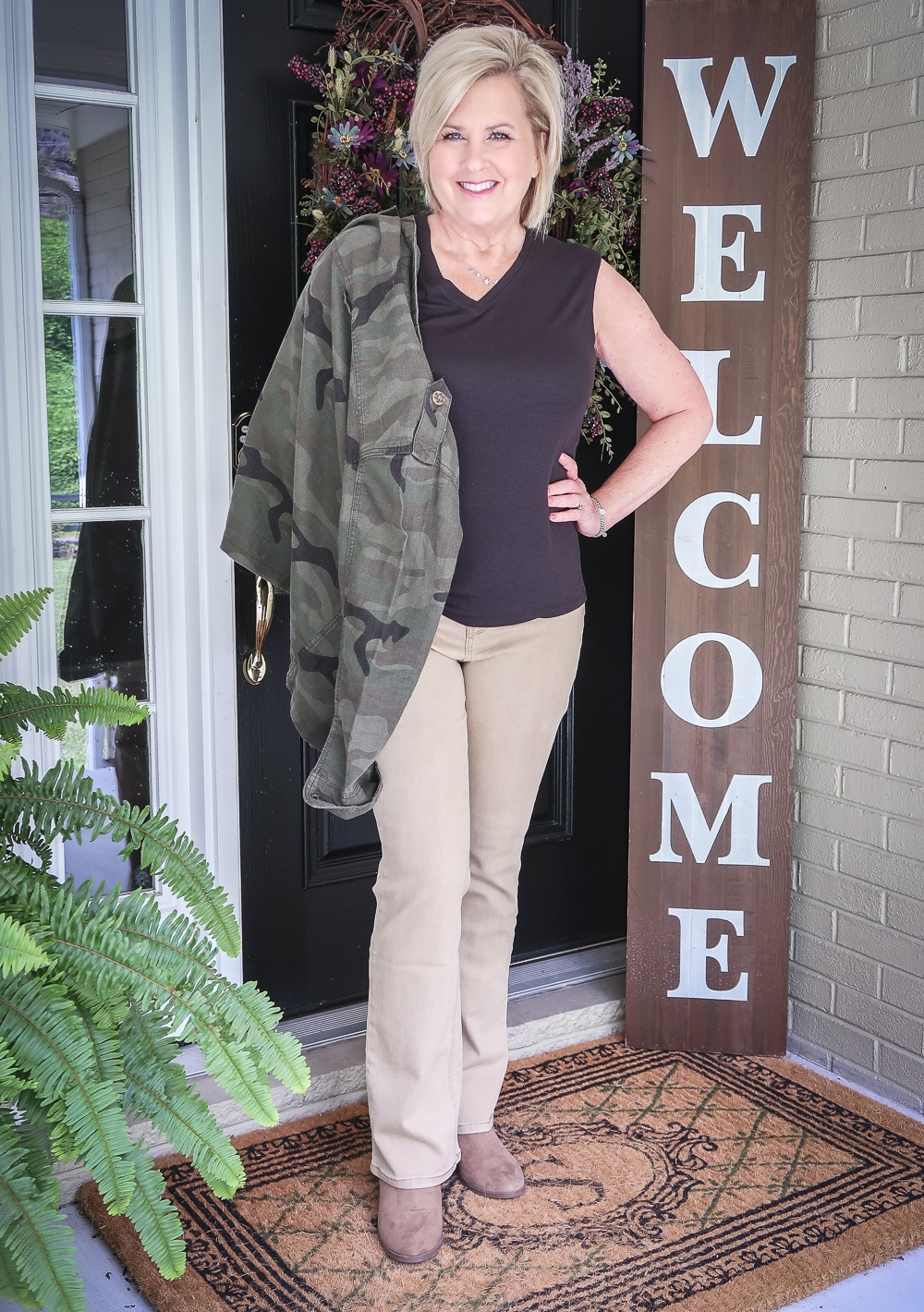 Fashion Blogger 50 Is Not Old is wearing a black v-neck tank top, beige bootcut jeans, taupe ankle booties, and carrying a olive green camo jacket all from Walmart