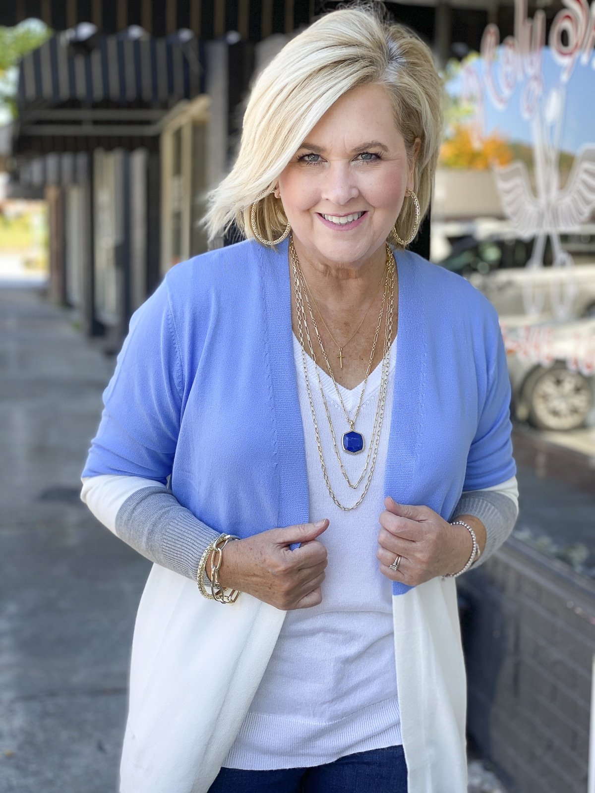 Fashion Blogger 50 Is Not Old is standing on a sidewalk in a color block sweater cardigan with gold jewelry