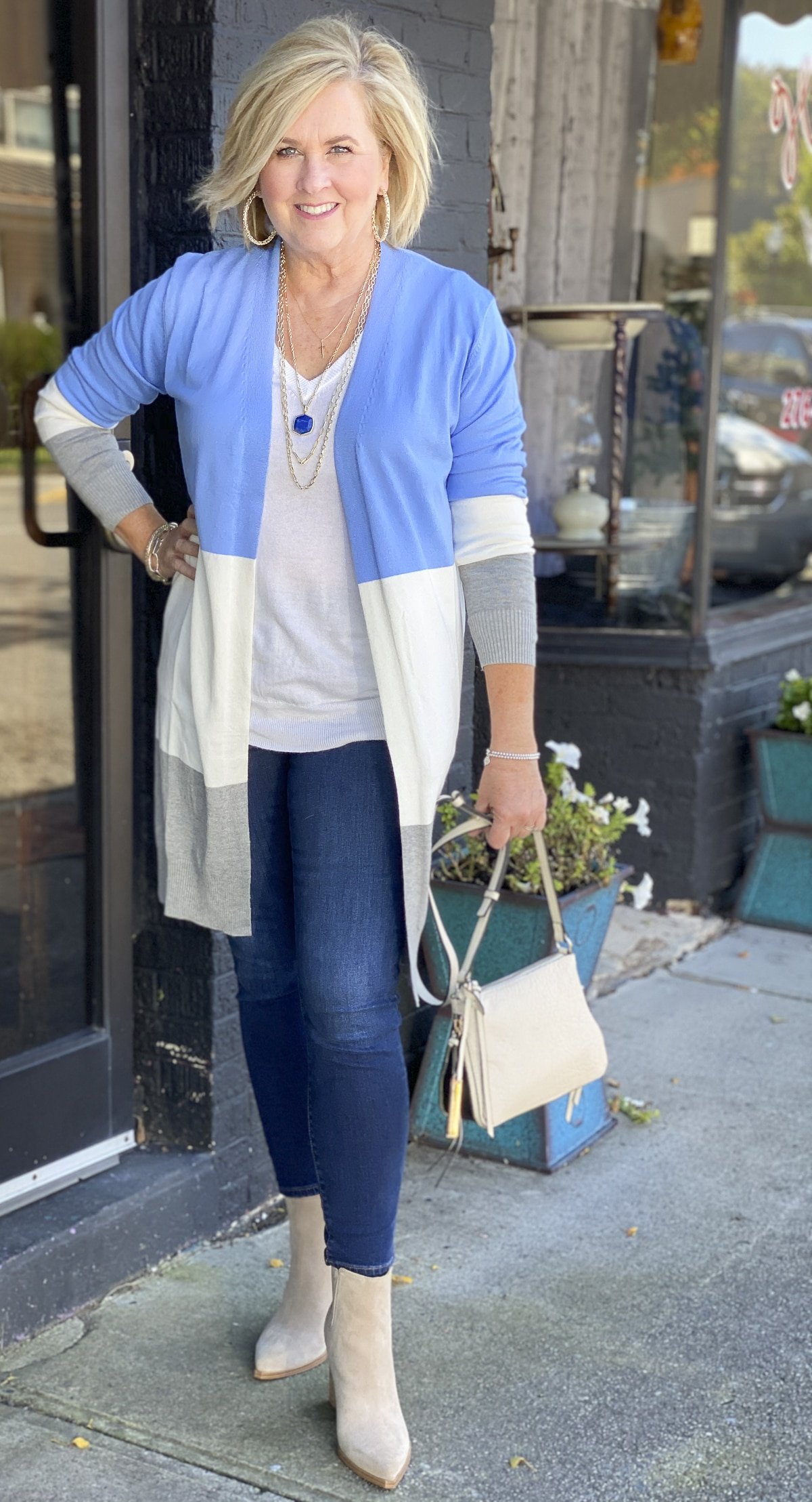 Fashion Blogger 50 Is Not Old is standing on a sidewalk in a color block sweater cardigan, dark skinny jeans, suede ankle boots, and holding a Vince Camuto crossbody handbag