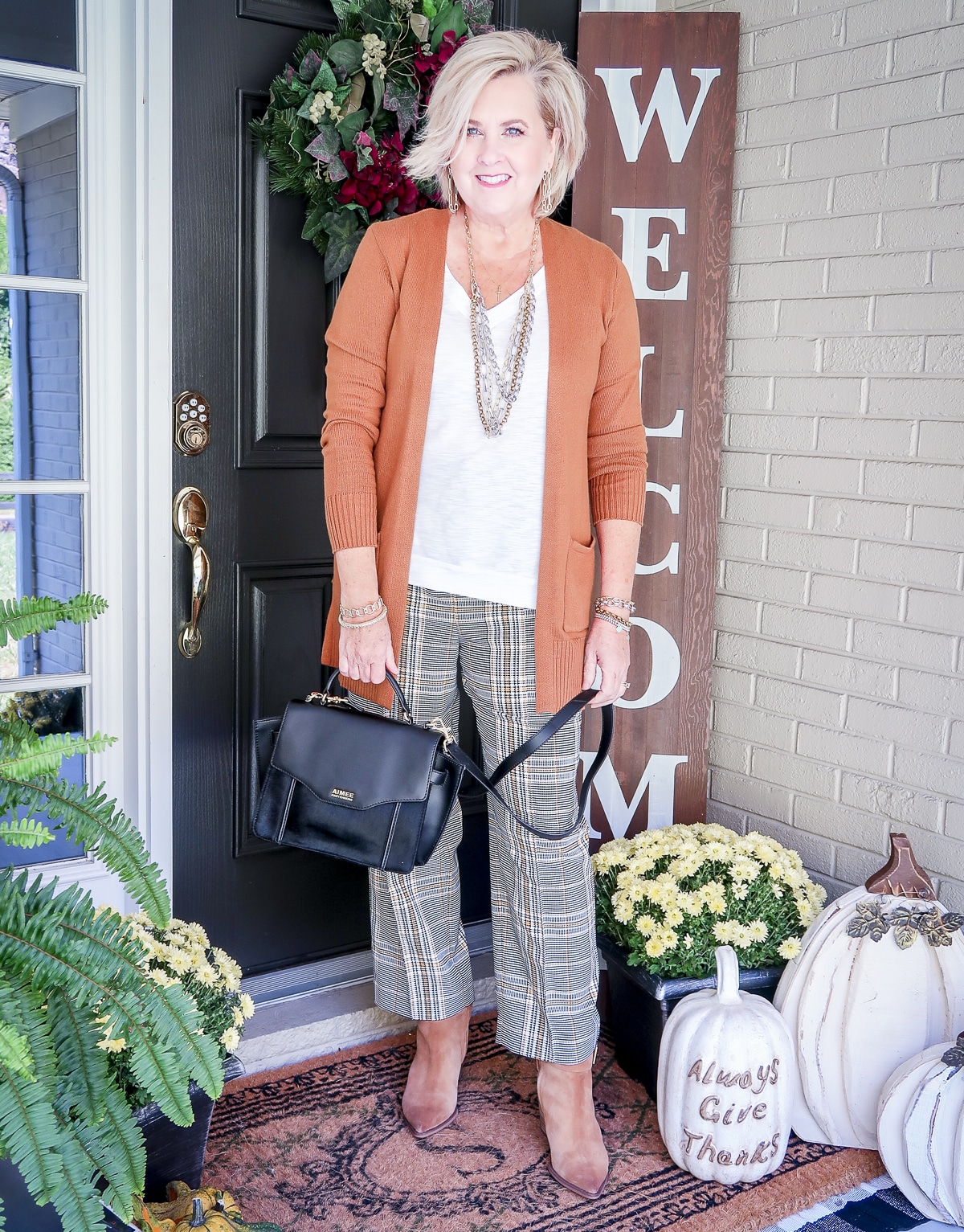 Fashion Blogger 50 Is Not Old is wearing a white tank with a caramel cardigan, plaid ankle pants, brown ankle boots, and carrying a black haircalf handbag