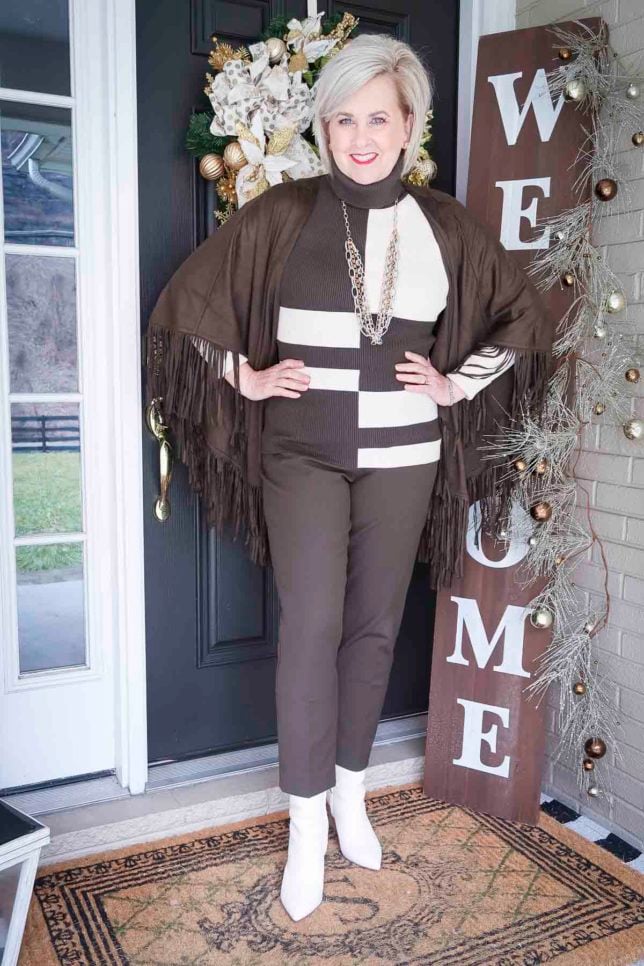Fashion Blogger 50 Is Not Old is wearing a colorblocked patterned turtleneck with ankle pants, a fringed ruana, and white ankle boots