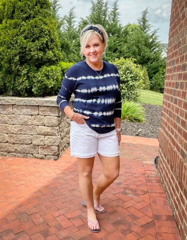 Fashion Blogger 50 Is Not Old is on a patio looking festive for the 4th of July in a blue and white tie-dyed sweater, a navy headband with stars, red gingham flip-flops, and white Bermuda shorts