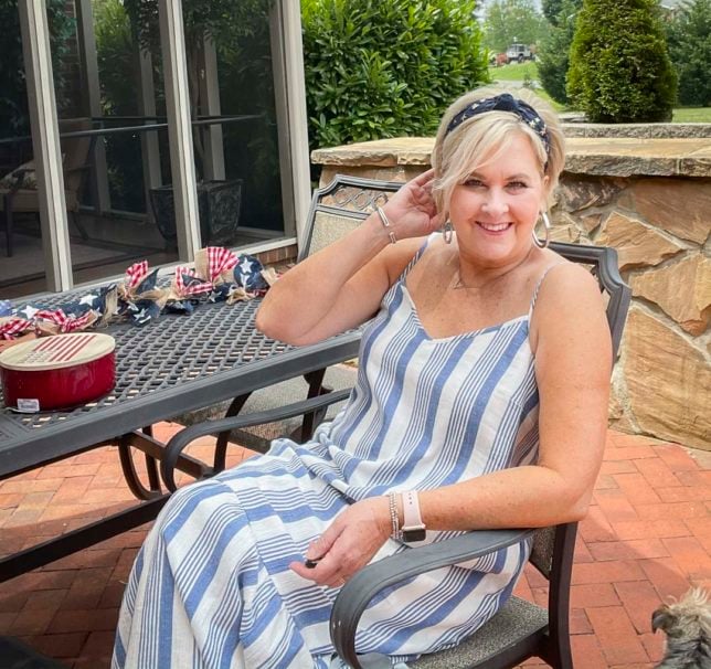 Fashion Blogger 50 Is Not Old is sitting at a patio table looking festive for the 4th of July in a blue and white striped sundress and a navy headband with stars