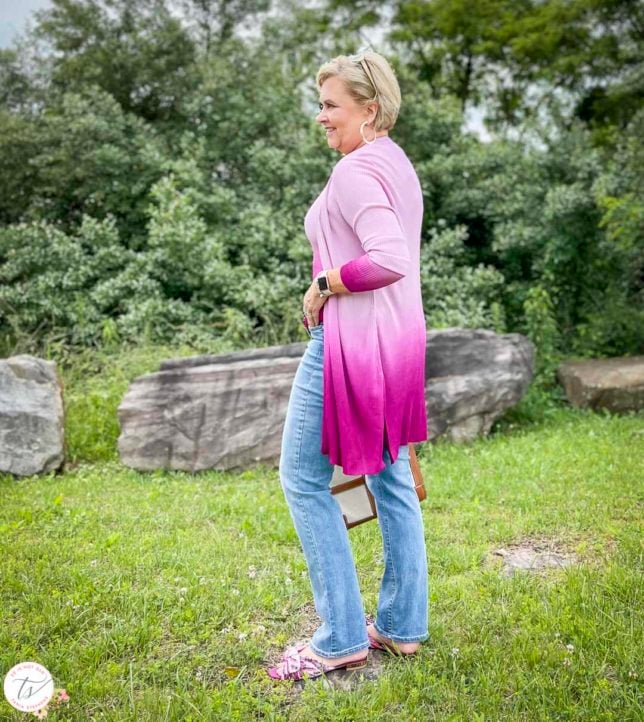 Fashion Blogger 50 Is Not Old is looking colorful in a pink ombre coverup, bootcut jeans, pink floral slides, and is carrying a Tory Burch satchel