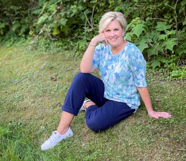 Fashion Blogger 50 Is Not Old is sitting on the ground in floral sweater, navy pants, and white sneakers from Appleseeds