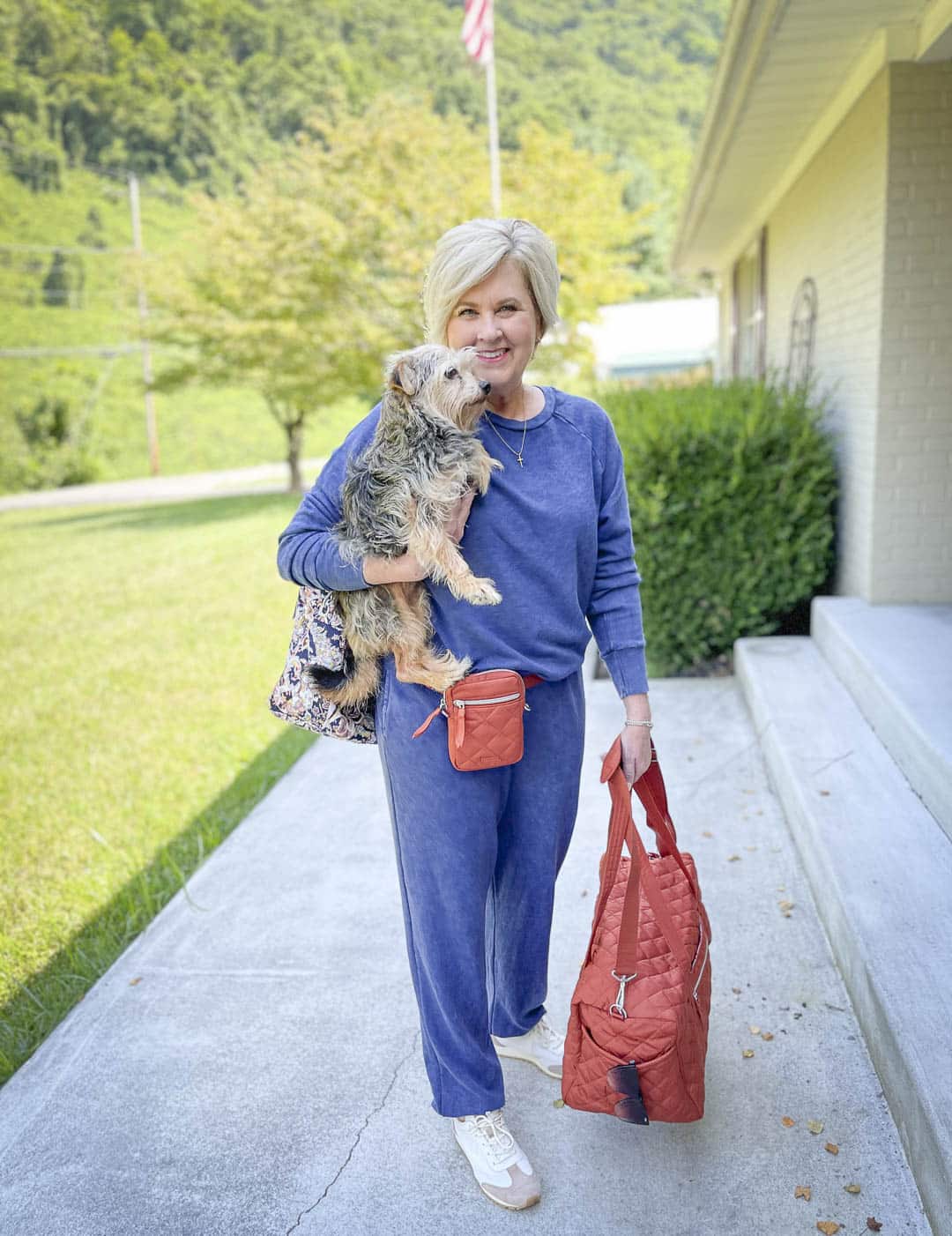 Over 40 Fashion Blogger, Tania Stephens, is holding her dog while wearing a navy sweatshirt and sweatpants set with Tory Burch sneakers and is all packed to go with her Vera Bradley weekender bags and Terracotta crossbody bag