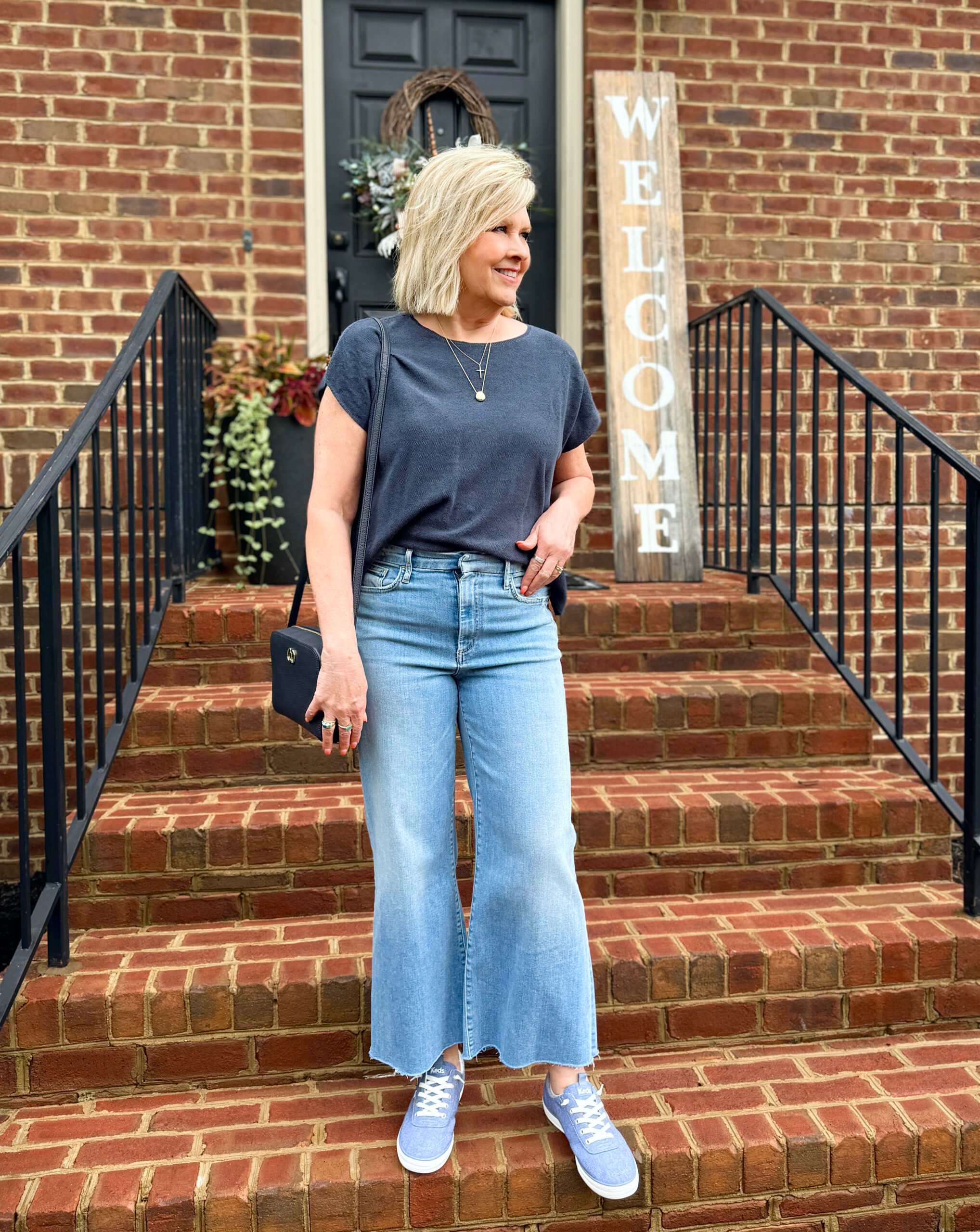 Woman in navy tee, light denim jeans, and chambray Keds standing on outdoor steps.