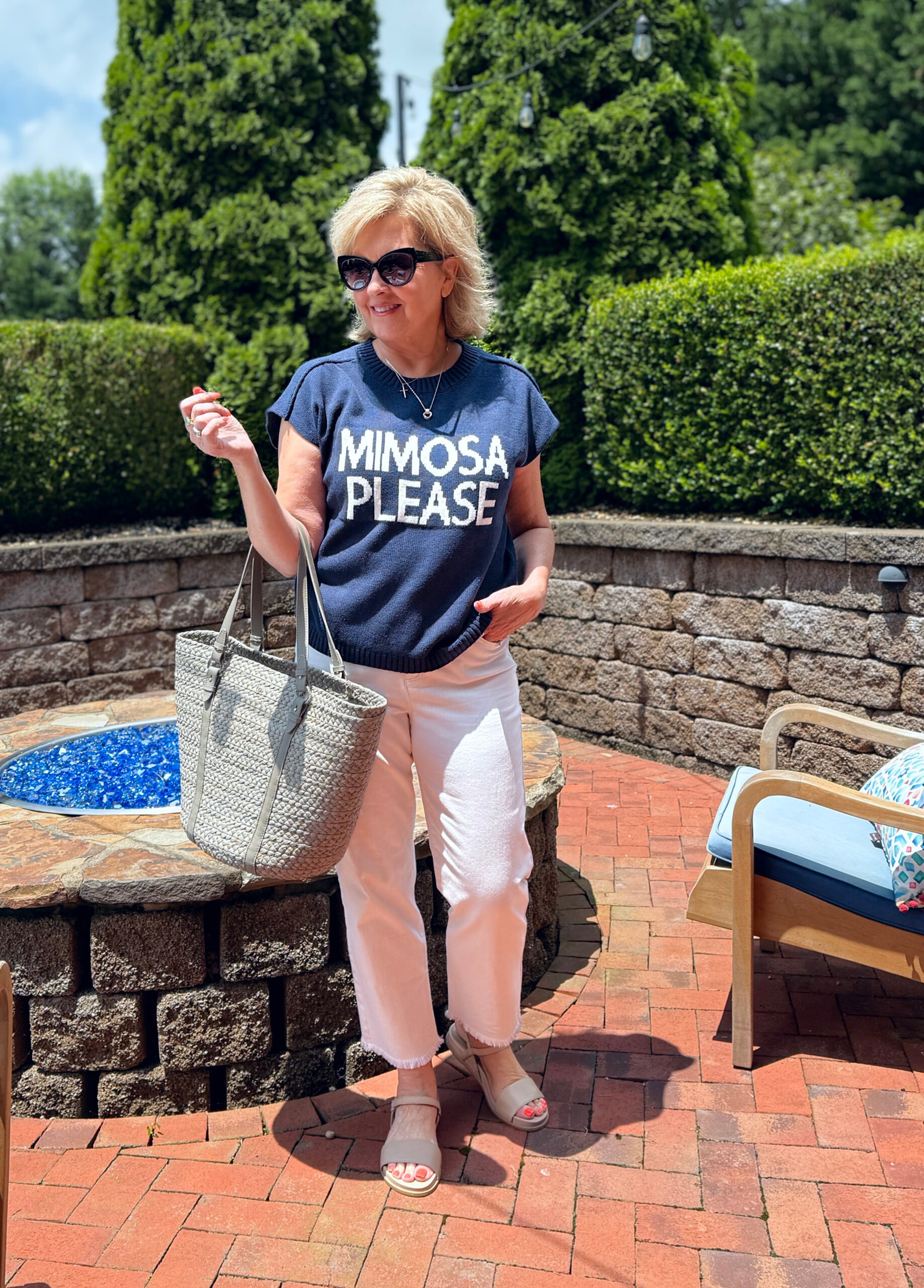 Woman in soft cap sleeve sweater, ankle jeans, khaki sandals, cat eye sunglasses, tan tote; smiling outdoors in bright sunlight.