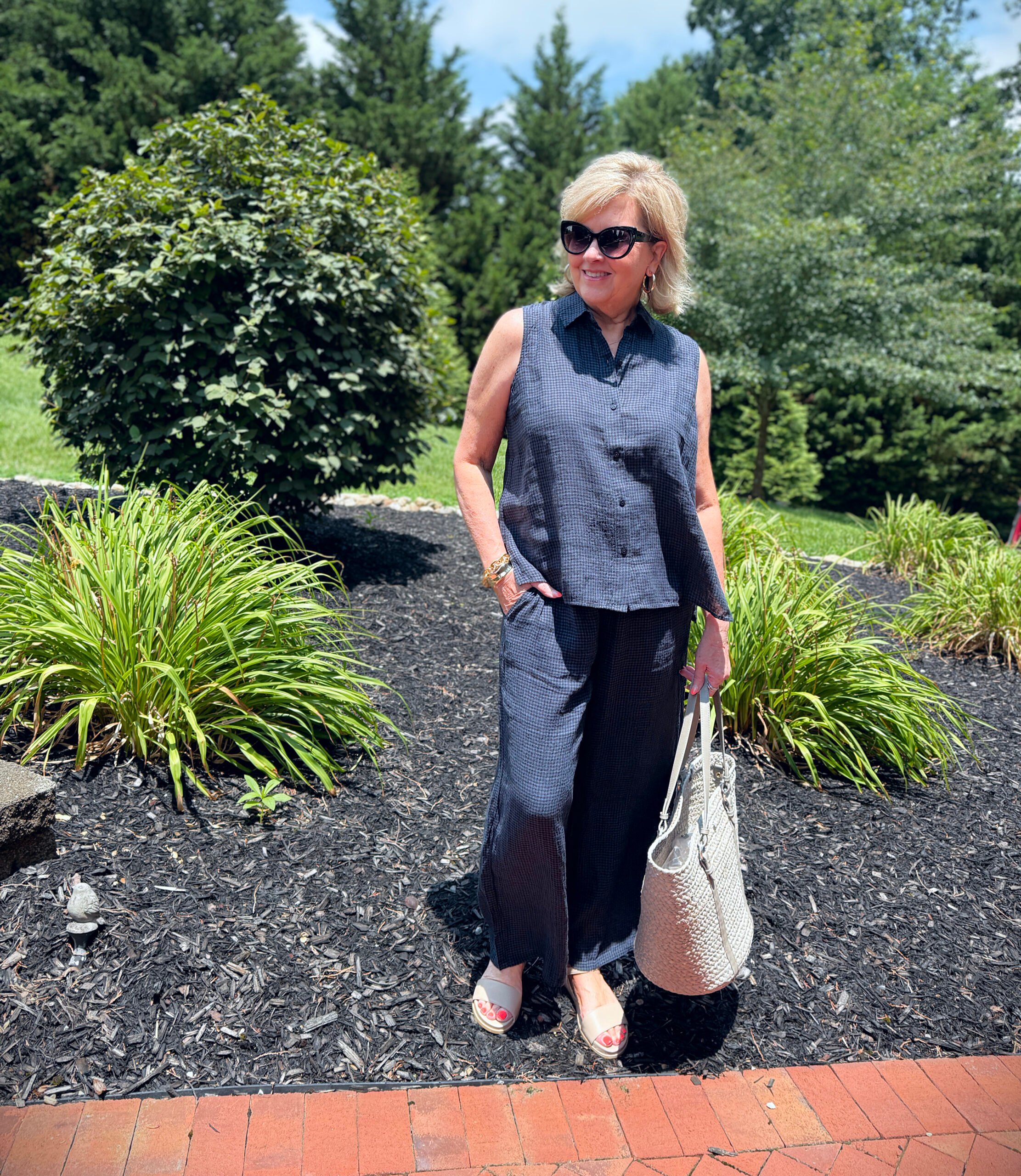 Woman wearing navy linen button-up with wide-leg linen pants, khaki sandals, cat eye sunglasses, tan tote; walking through a garden path.