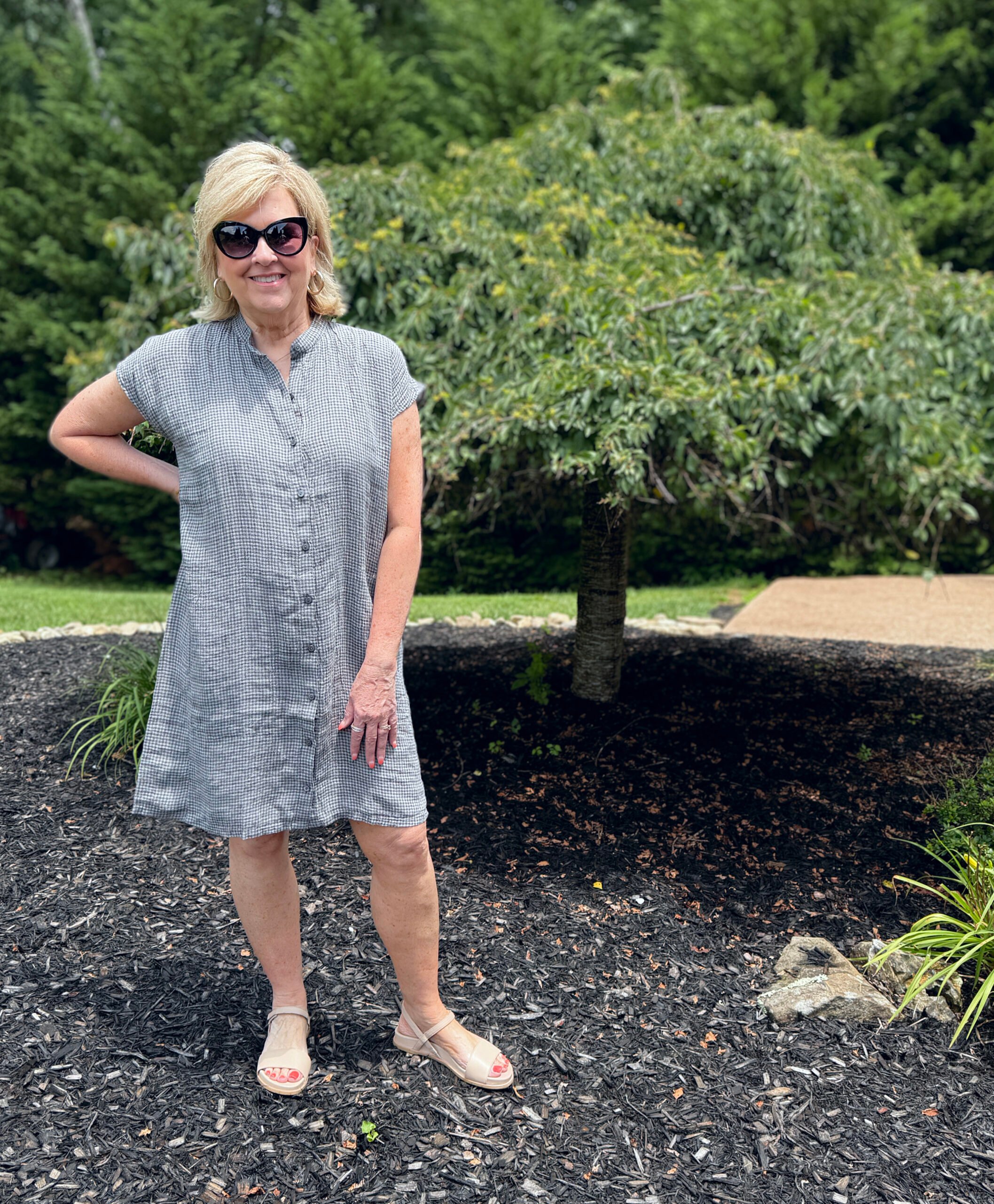 Woman in gingham linen shirtdress, khaki sandals, cat eye sunglasses; sitting at a park bench.