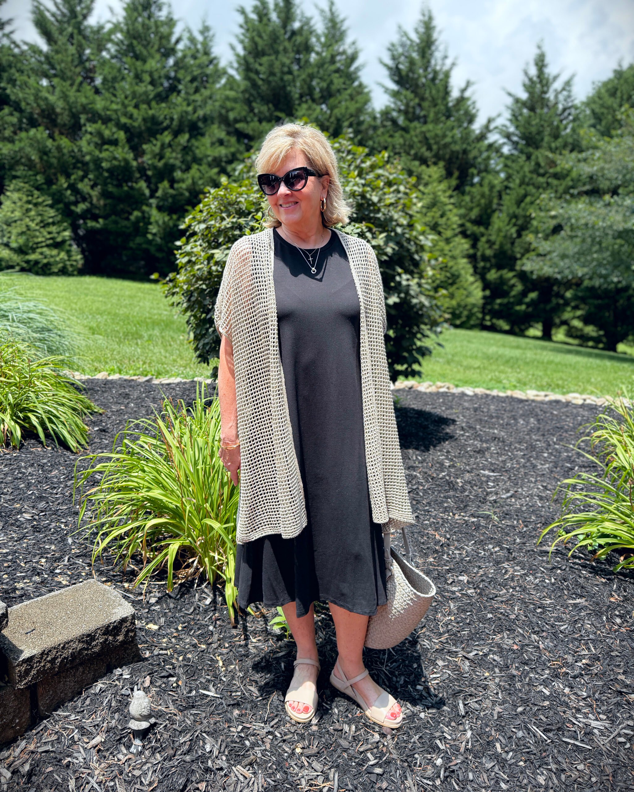 Woman wearing navy jersey shift dress with longline linen cardigan, khaki sandals, tan tote; leaning on a porch railing.
