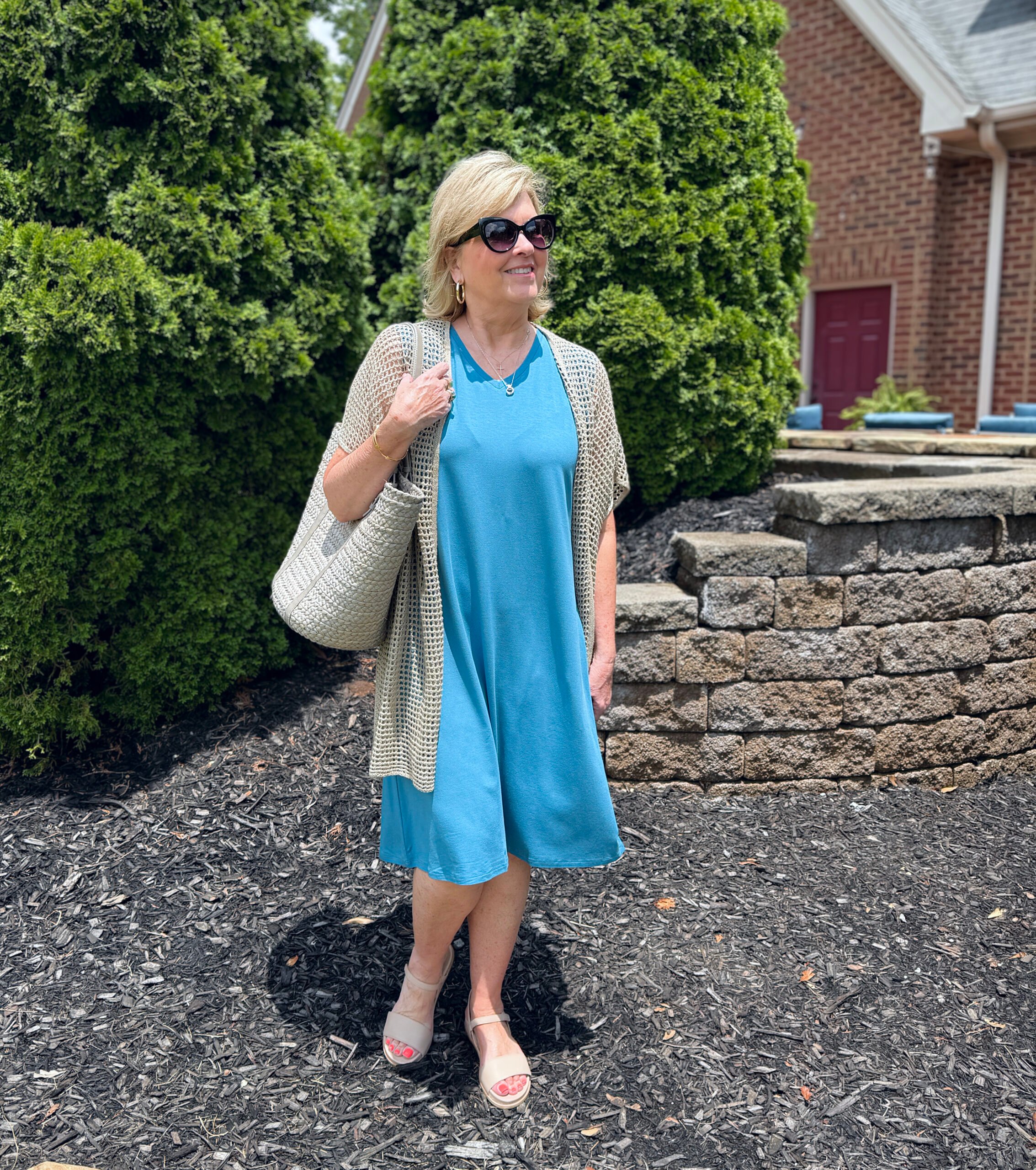 Woman in marina blue V-neck tank dress with linen cardigan, khaki sandals, cat eye sunglasses, tan tote; strolling along a flower-lined walkway.