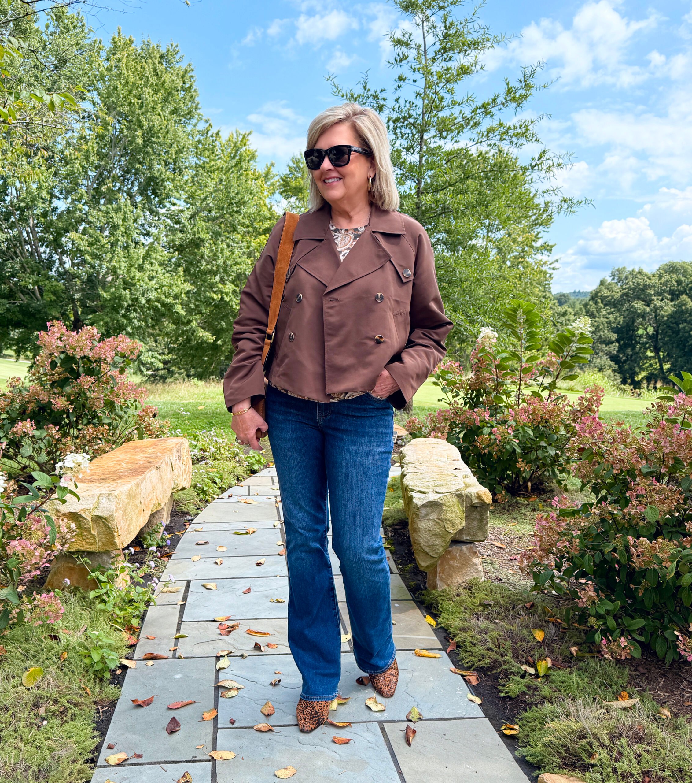 Woman wearing a Steve Madden short trench coat layered over a paisley print top with bootcut jeans and fall accessories.