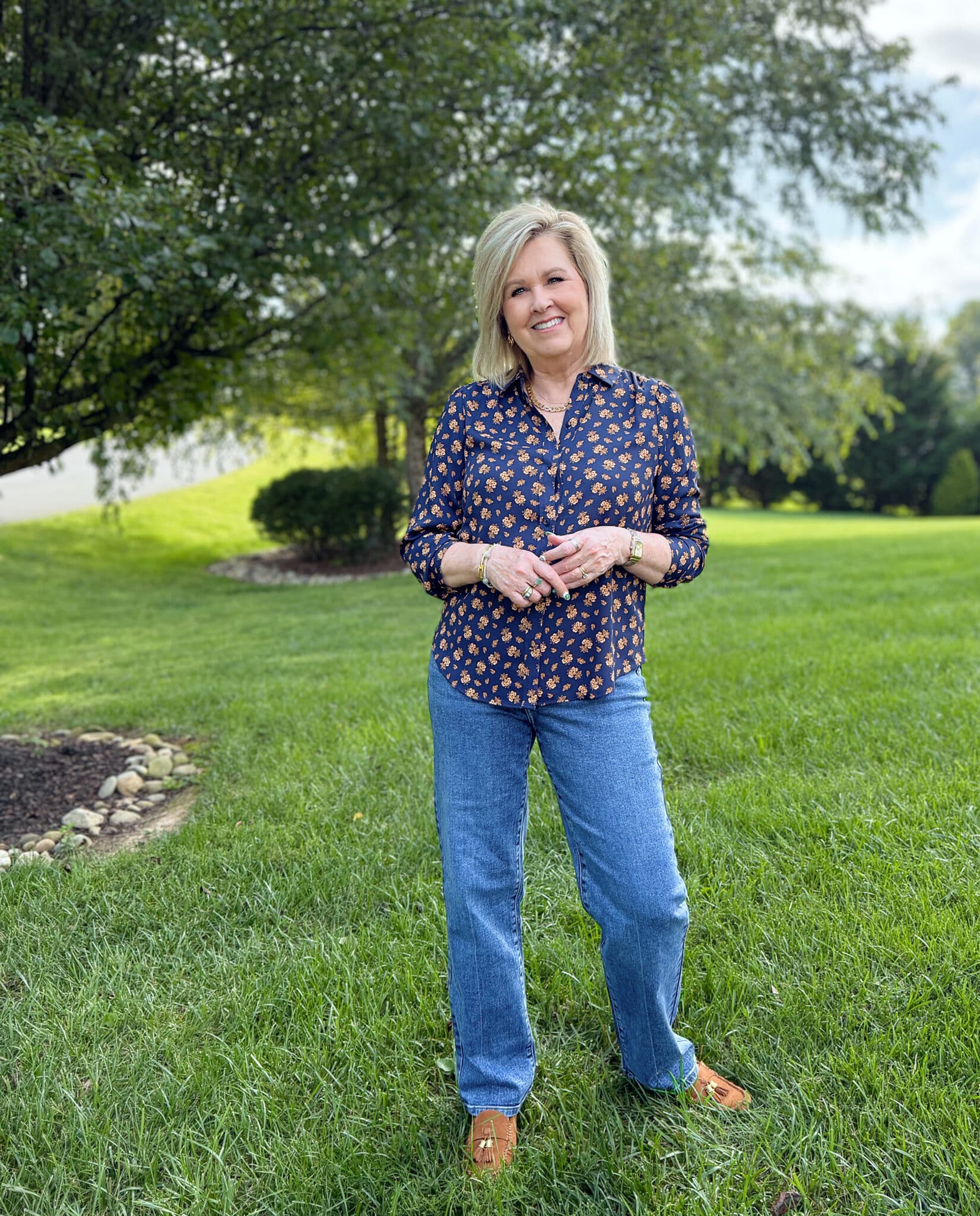 Woman over 50 standing in a grassy field in Kohl's straight leg jeans and a button floral shirt for fall