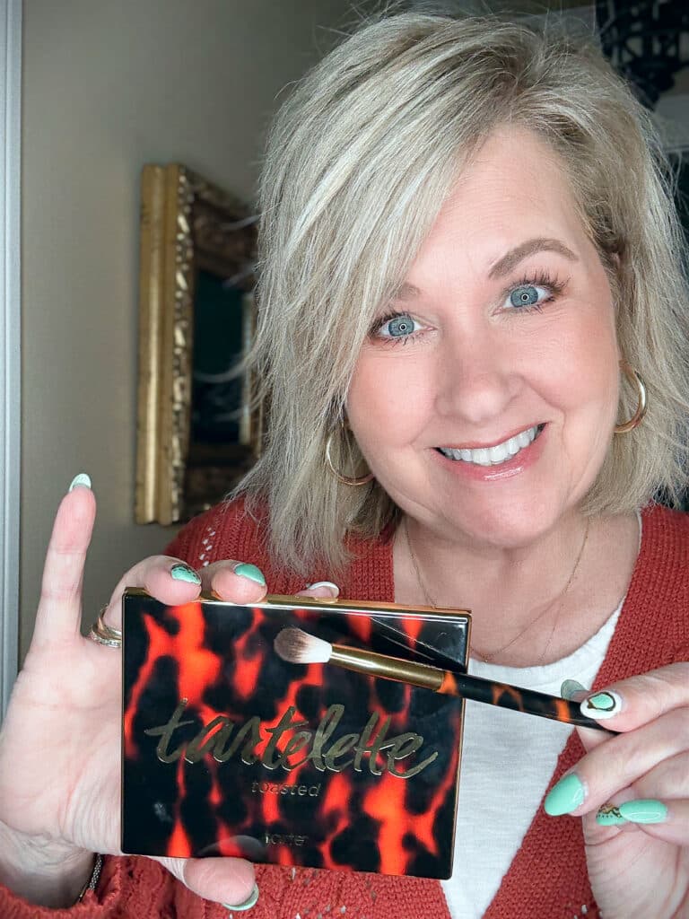 Tania in an orange sweater, holding a tarte Toasted Eyeshadow Palette and brush