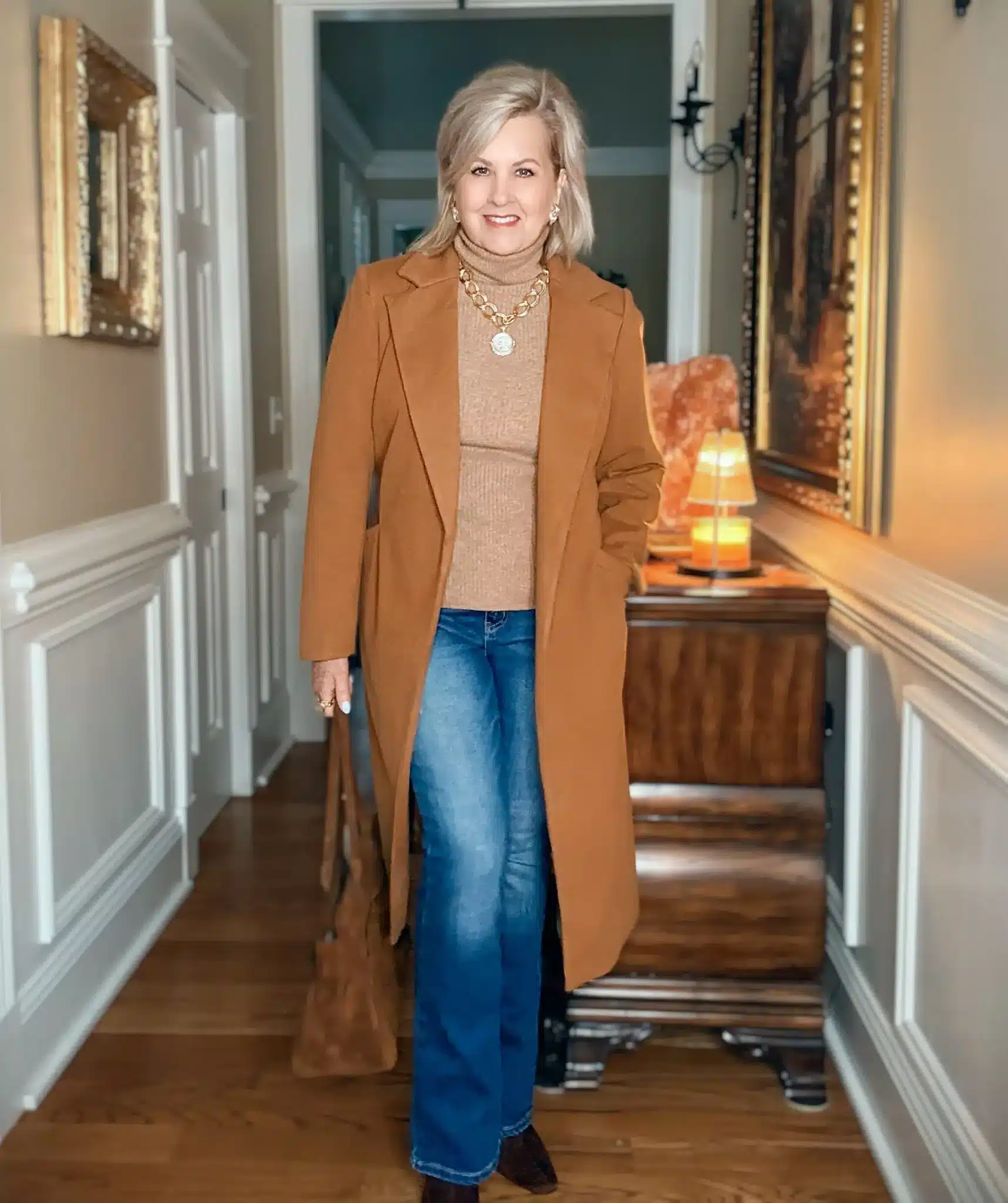 Tania wearing a camel coat and turtleneck with jeans, gold jewelry, and brown boots for a cozy Thanksgiving outfit.