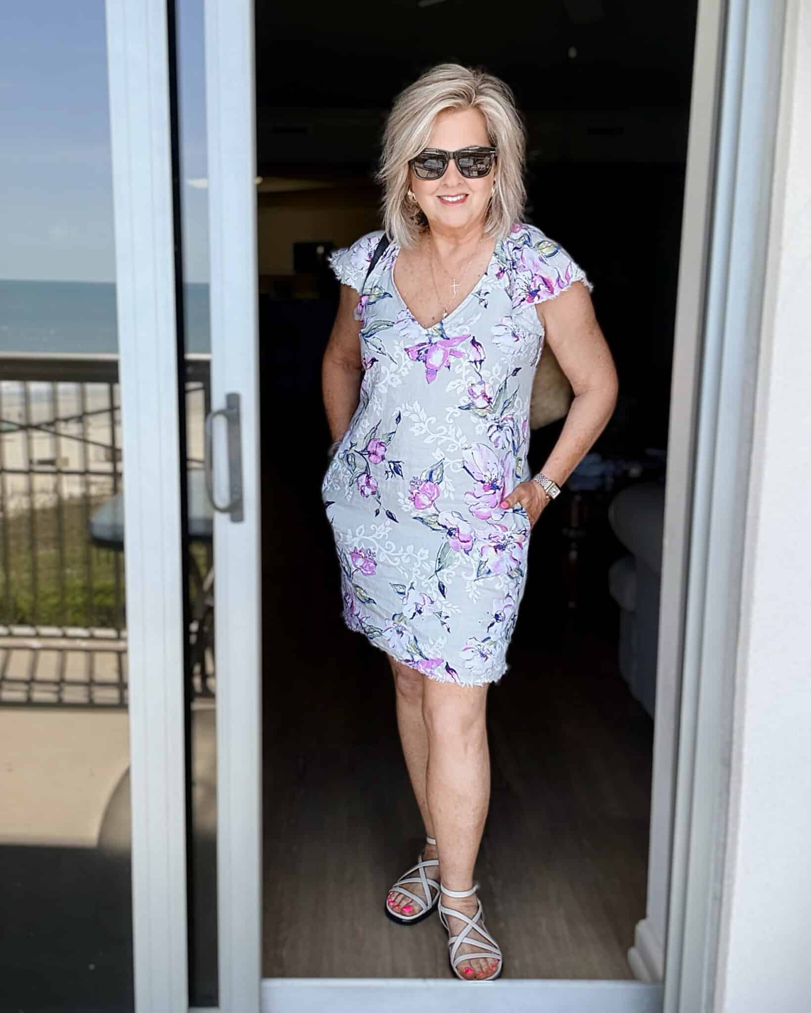 Woman over 50 wearing a light blue floral linen shift dress with silver sandals at the beach.