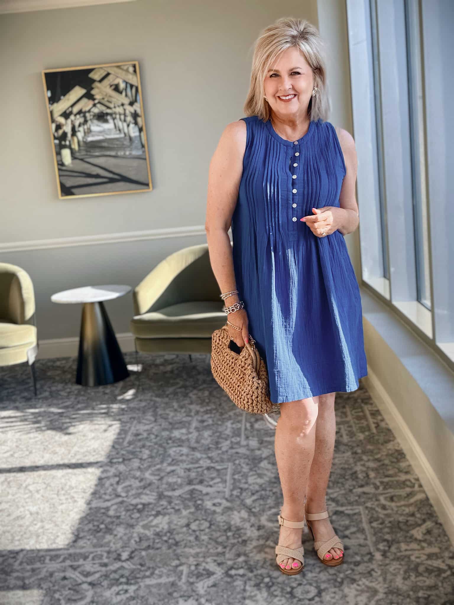 Woman over 50 wearing a blue organic cotton gauze sleeveless swing dress with nude heeled sandals and a raffia clutch.