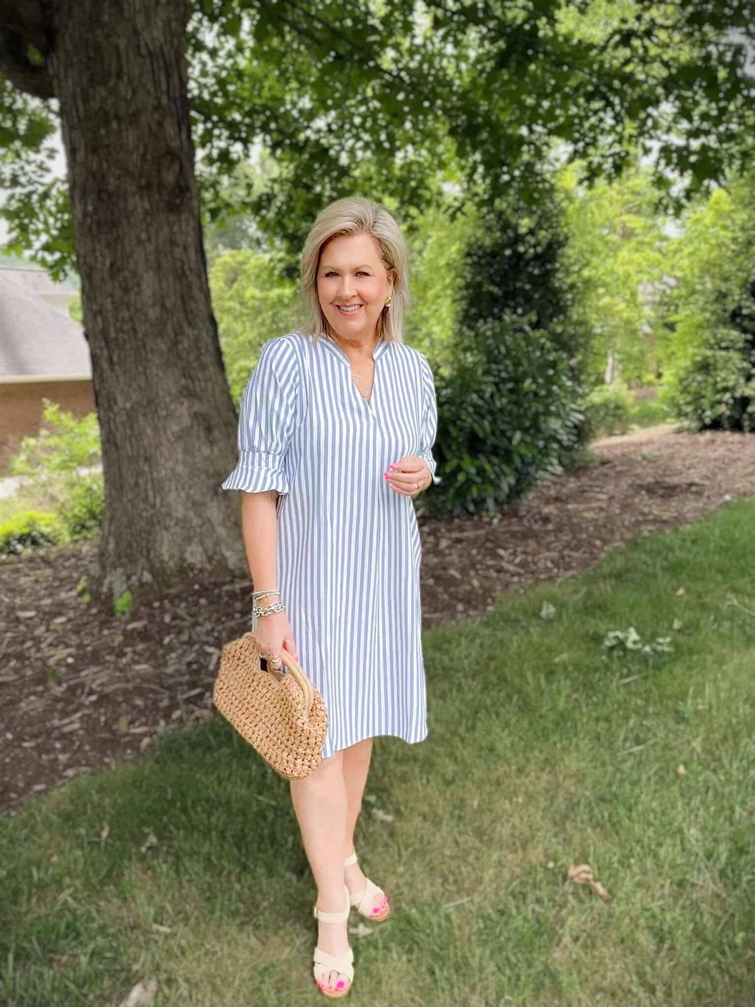 Woman over 50 wearing a blue and white striped shift dress with ruffle sleeves and a raffia handbag for summer.