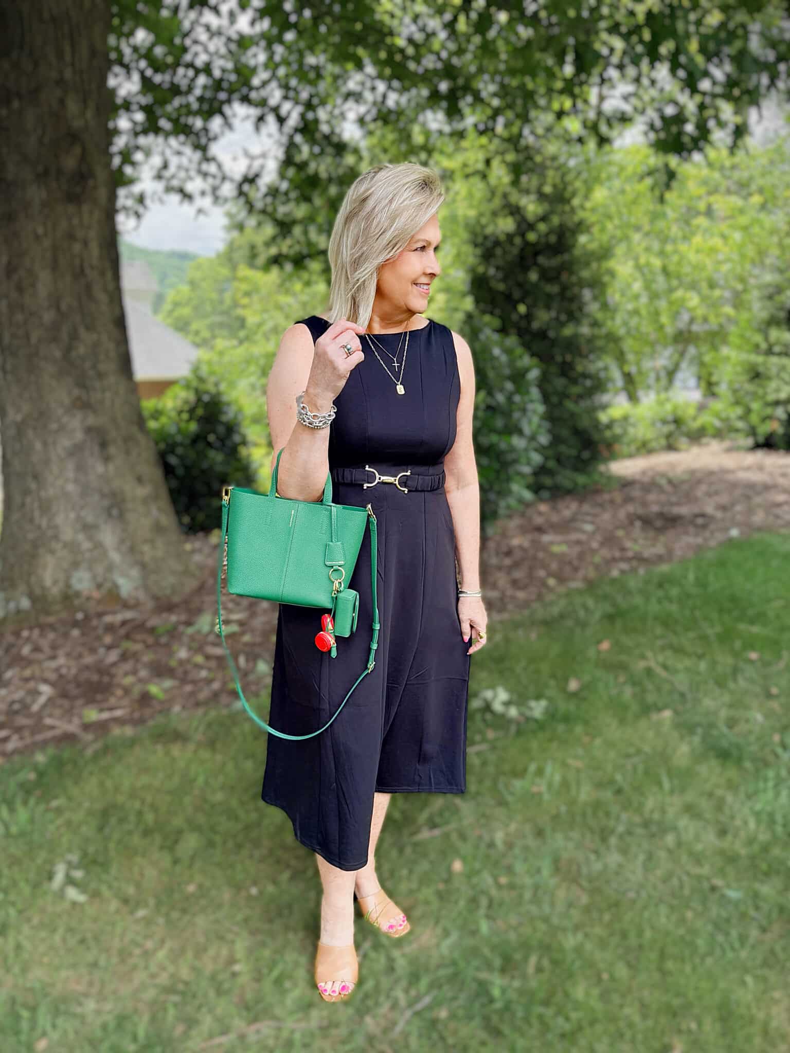 Woman over 50 wearing a sleeveless black midi dress with a green tote bag and tan sandals for a polished summer outfit.