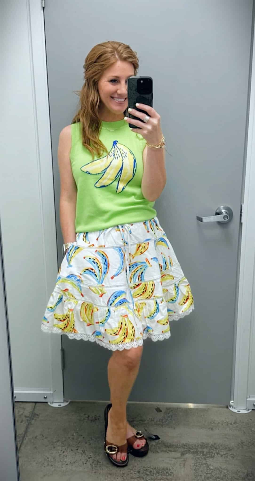 Stephanie wearing a lime green banana graphic sweater shell with a white tiered banana-print mini skirt and brown jelly sandals.